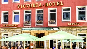 Exterior del Hotel Victoria con letreros de restaurante y bistró, y mesas al aire libre con sombrillas.