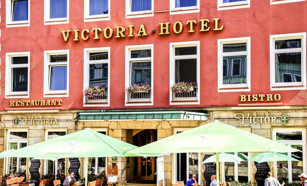 Exterior del Hotel Victoria con letreros de restaurante y bistró, y mesas al aire libre con sombrillas.