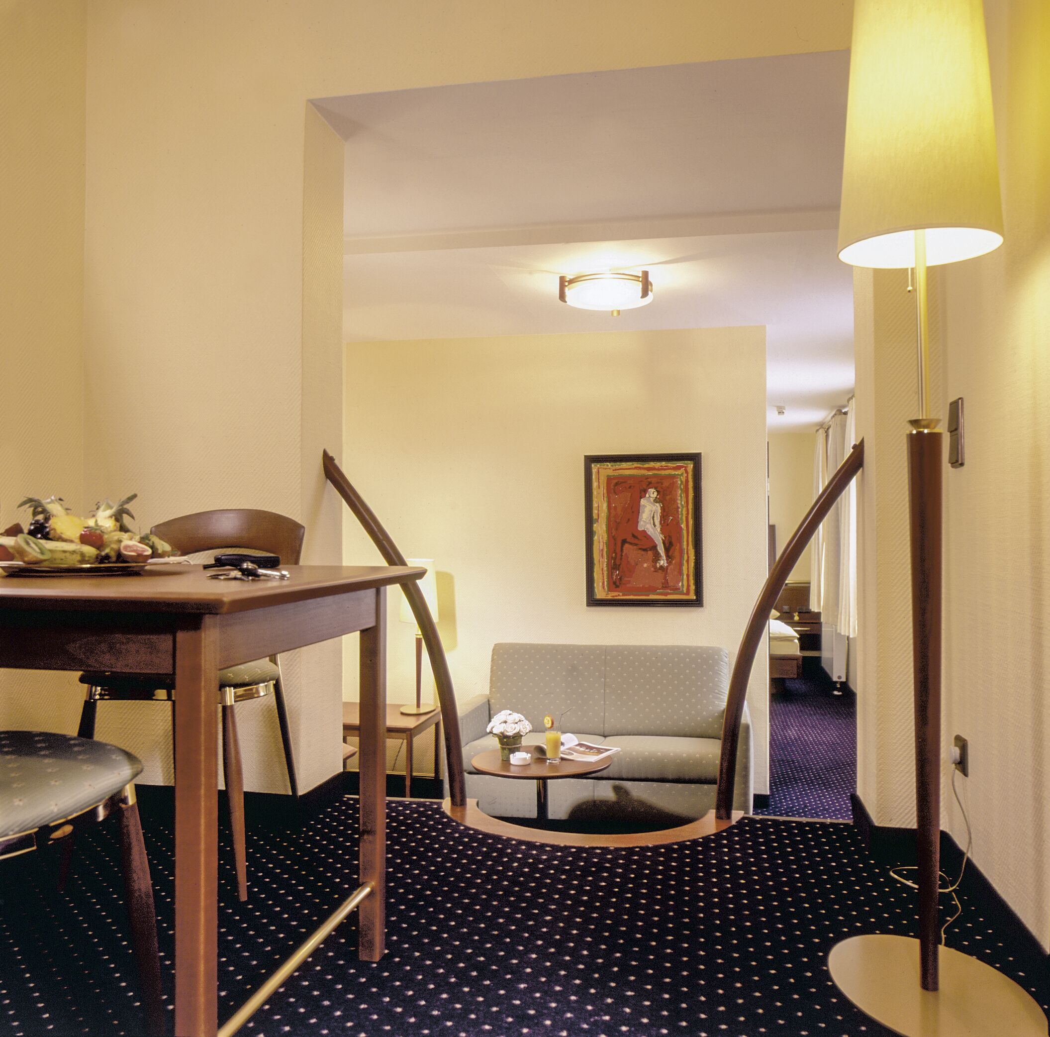 A hotel room with a dining table, a sofa, a small table with a vase of flowers, and a painting on the wall.