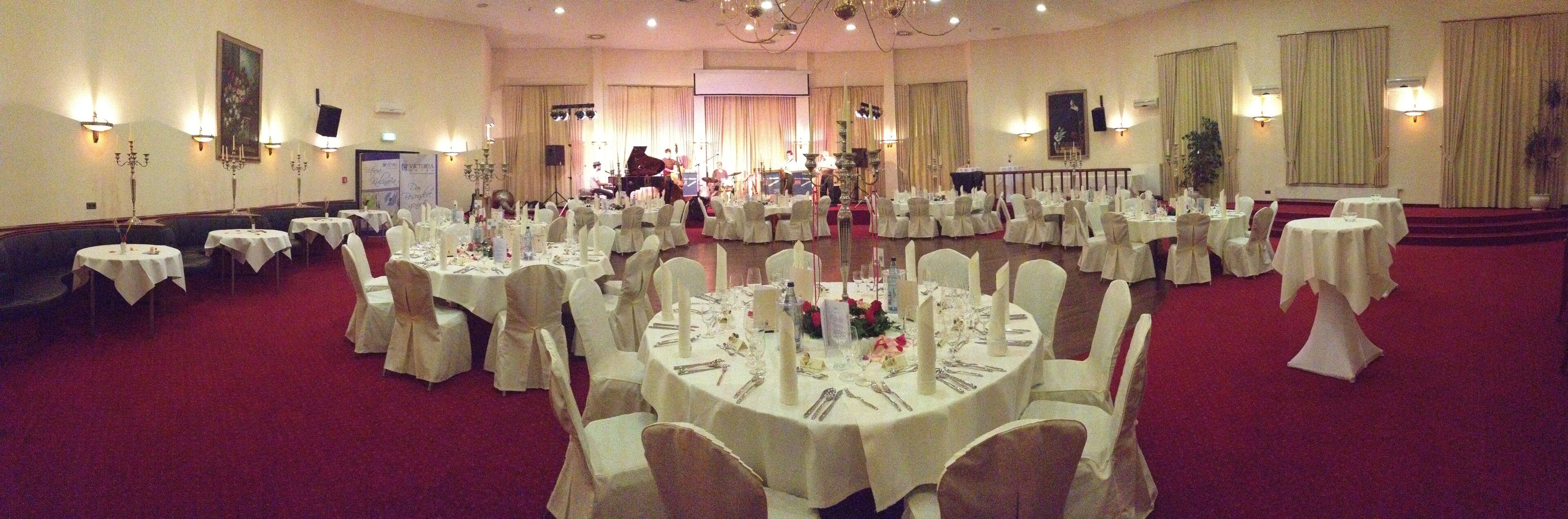 A banquet hall with round tables set for a formal event, a band performing in the background, and a stage with a piano.