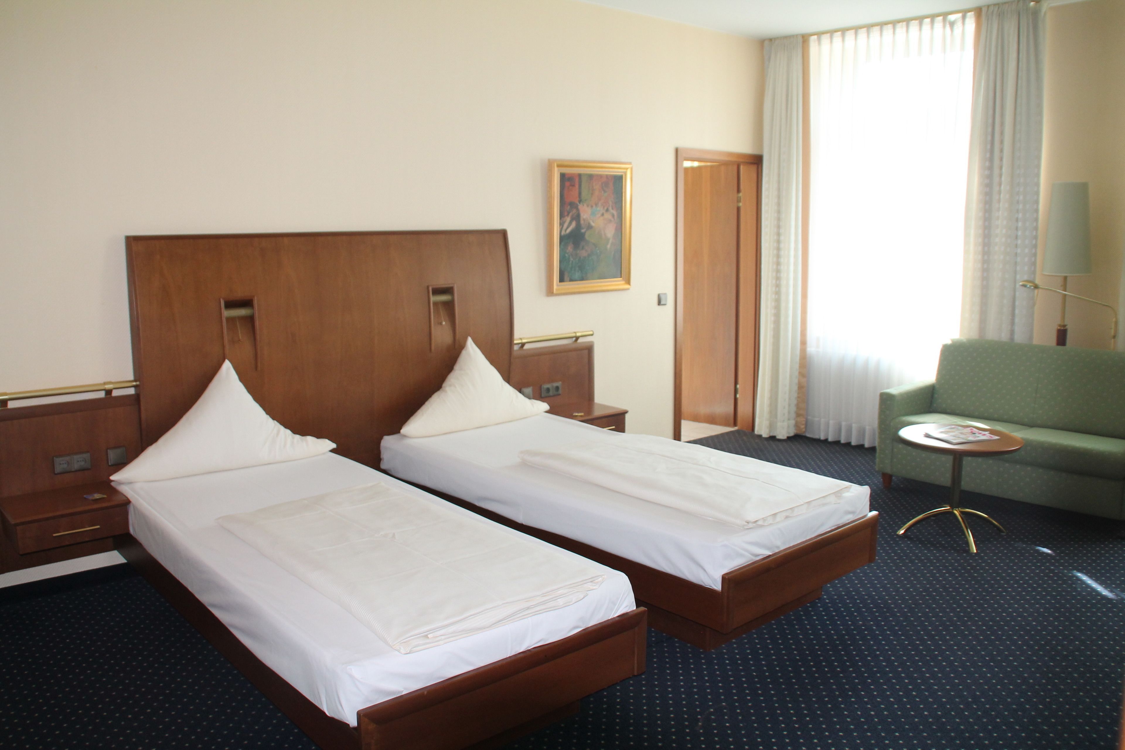 Een hotelkamer met twee eenpersoonsbedden, een groene fauteuil, een klein rond tafeltje en een schilderij aan de muur.
