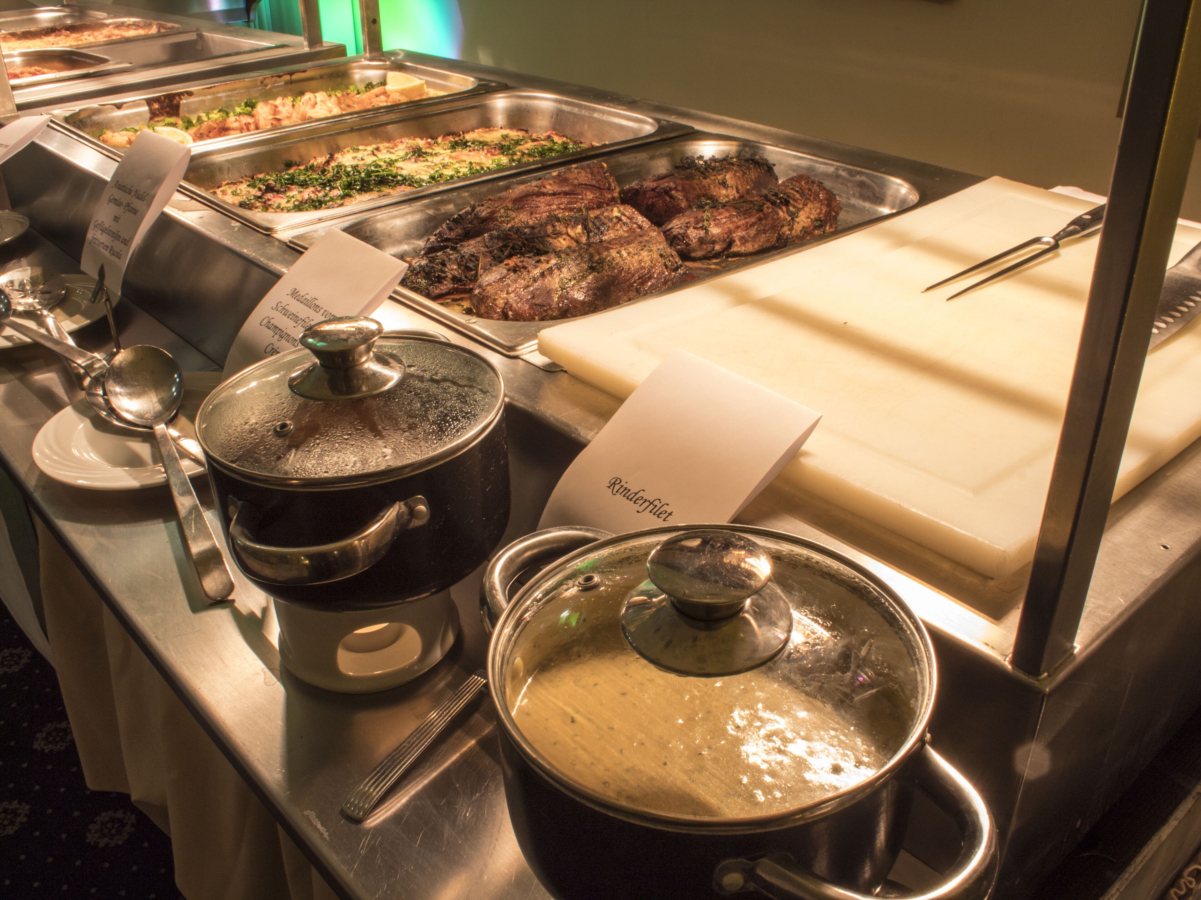 Ein Buffet mit verschiedenen Speisen, darunter eine Suppentopf, ein Schneidebrett und ein Schild mit der Aufschrift 'Rinderfilet'.