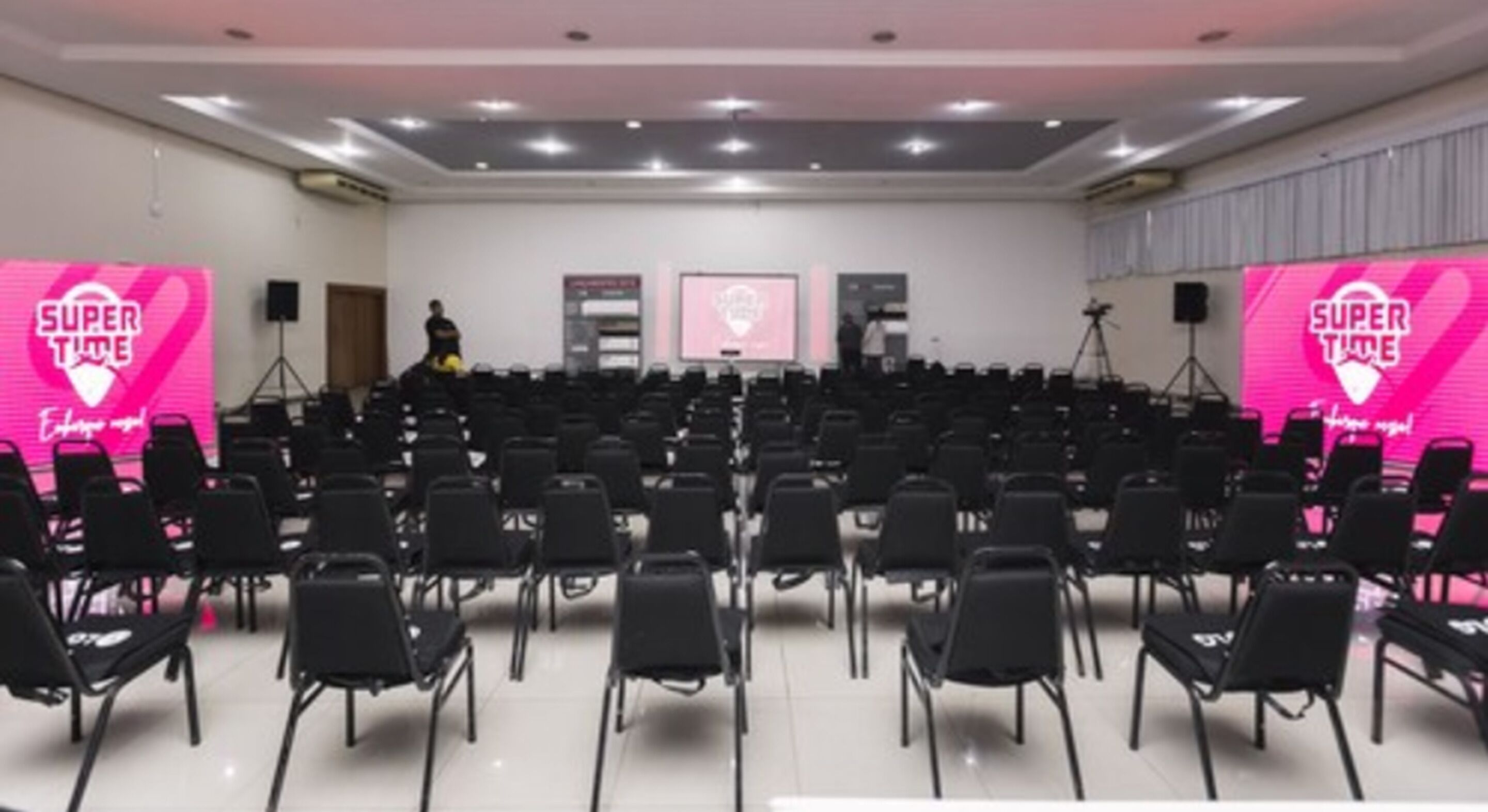 Un salón de conferencias con filas de sillas, un escenario con fondo rosa y una pantalla grande.