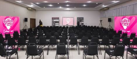 Un salón de conferencias con filas de sillas, un escenario con fondo rosa y una pantalla grande.