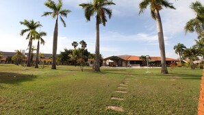 A grassy area with palm trees and a building with a red roof in the background. - Ipê Park Hotel (São José do Rio Preto)
