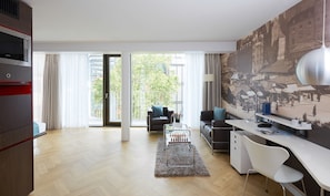 A modern living room with a sofa, a coffee table, and a desk with a lamp. There is a large window with curtains and a view of buildings outside. - Living Hotel Das Viktualienmarkt (Munich)