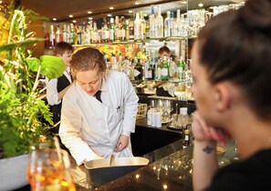 Un coin bar avec un barman préparant une boisson, des étagères remplies de bouteilles et un client assis au bar.