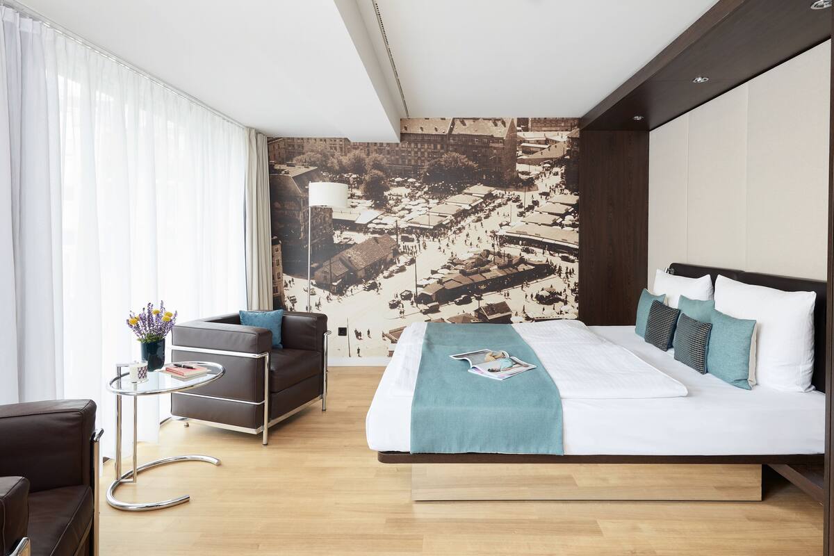 A modern hotel room with a large bed, a sofa, a small table, and a view of a cityscape through the window.