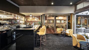Eine Bar mit Barhocker, einer Theke und Regalen voller Gläser und Flaschen. Im Essbereich stehen Stühle und Tische bereit, und im Hintergrund ist ein großes Fenster zu sehen.