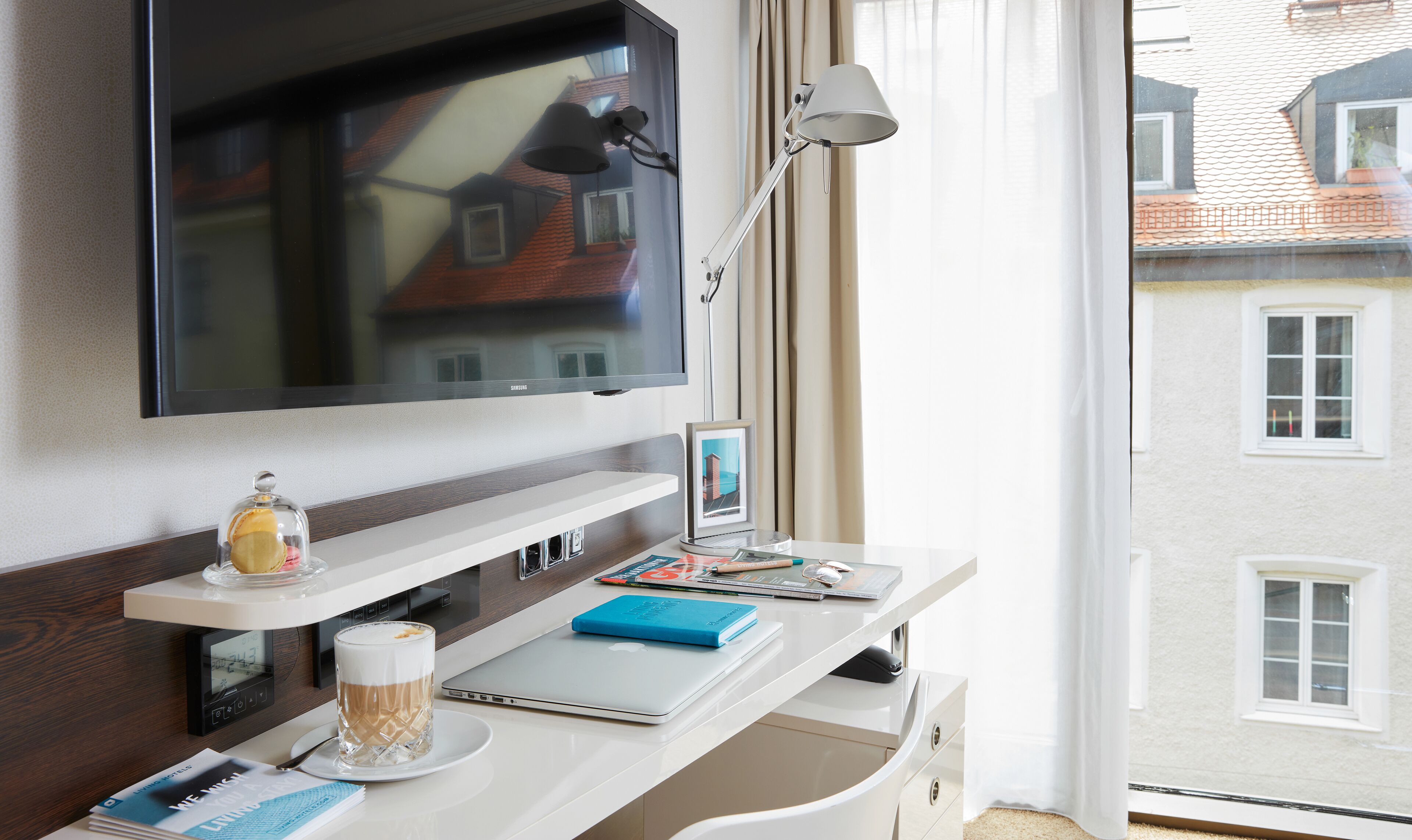 Habitación de hotel con televisión de pantalla plana en la pared, un escritorio con una computadora portátil, una taza de café y un cuadro enmarcado.