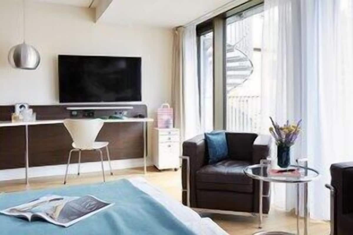 A modern hotel room with a bed, a desk, a TV, a sofa, and a small table with a vase.