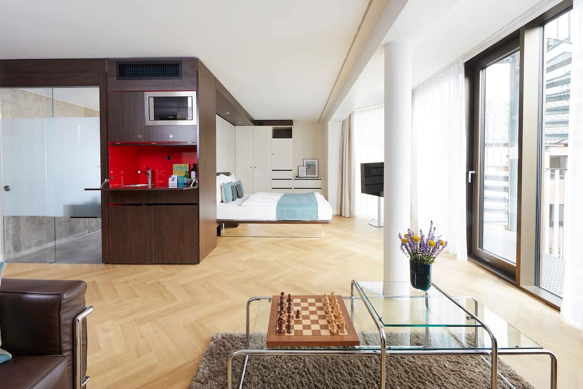 A modern hotel room with a kitchenette, a bed, a glass coffee table with a chessboard, and a view of the city through large windows.