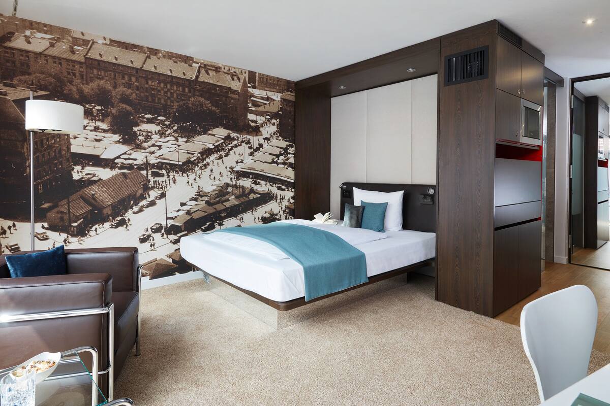 A modern hotel room with a large bed, a desk, and a view of a cityscape through the window.