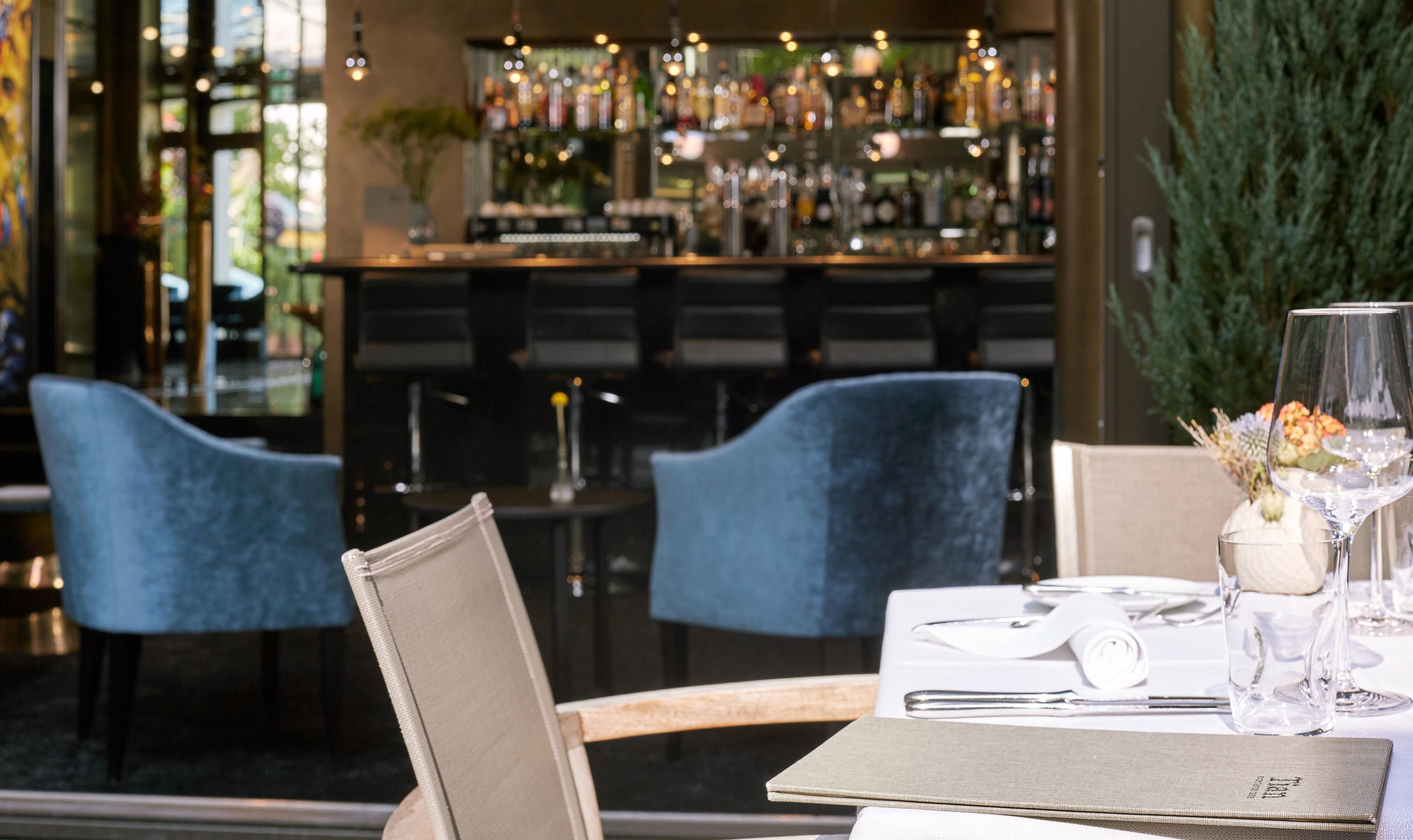 Interior de restaurant amb zona de bar al fons, mostrant diverses ampolles i una planta en test.