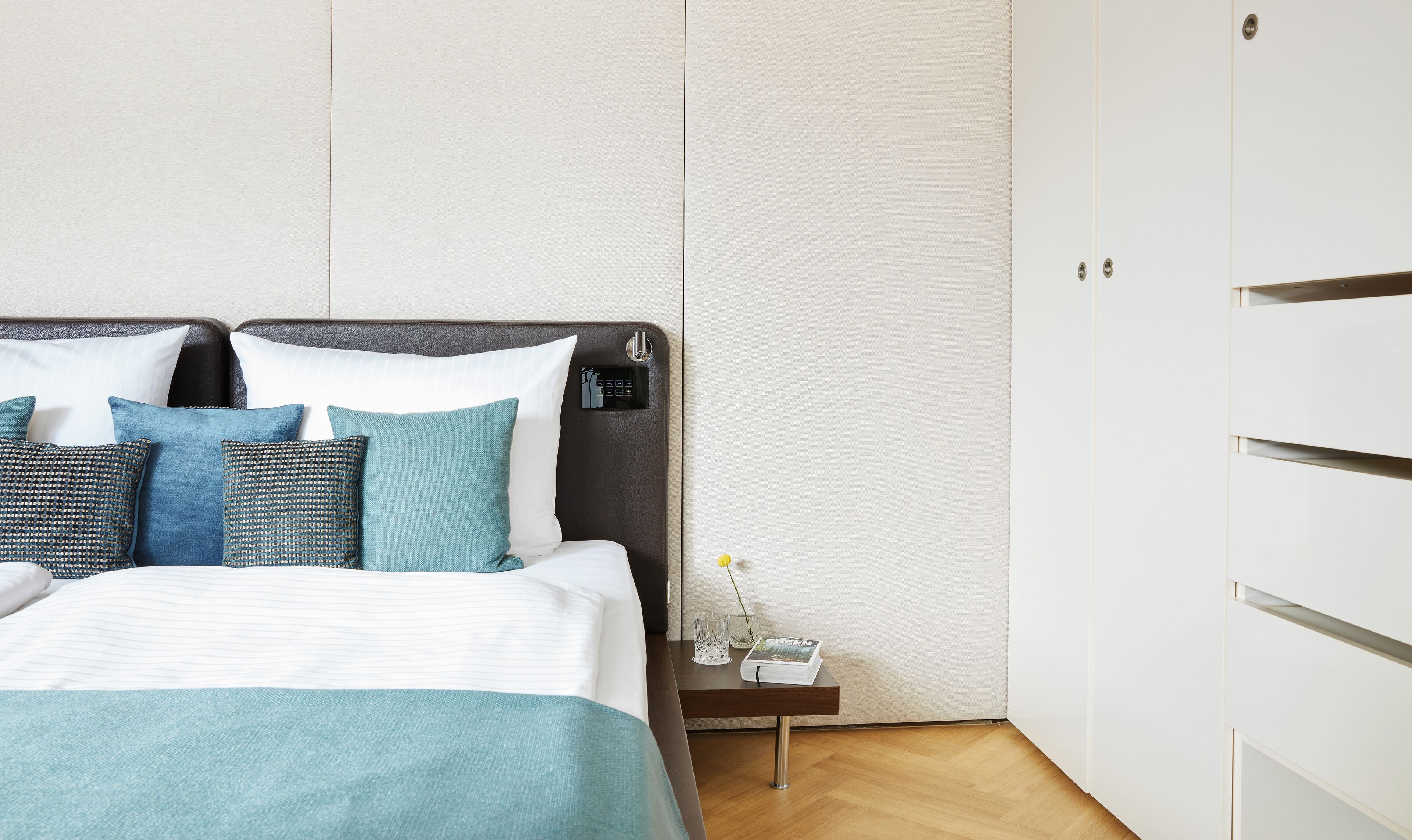 Een keurig opgemaakt bed met een donkere hoofdbord, een nachtkastje met een boek en een vaas, en een garderobe met een spiegel.