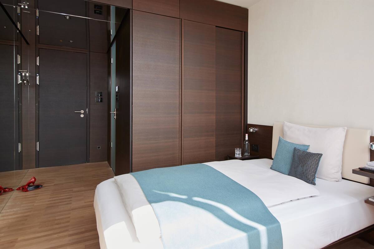 A hotel room with a large bed, a wooden wardrobe, a bedside table with a lamp, and a chair.
