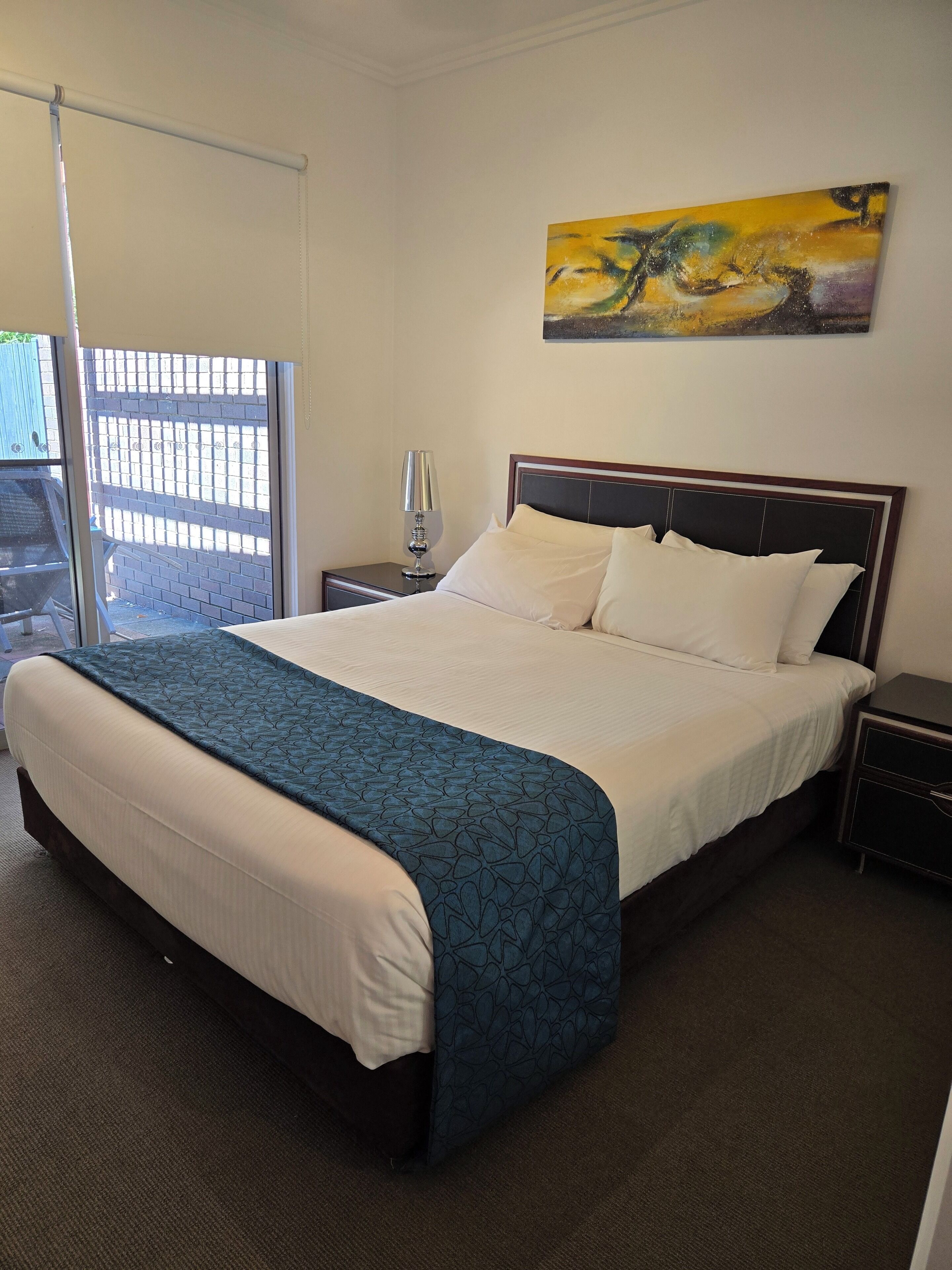 A neatly made bed with a blue patterned bedspread, a white pillow, and a wooden headboard. There is a painting on the wall above the bed, a bedside lamp on the nightstand, and a view of the outside through a window.
