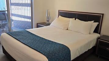 A neatly made bed with a blue patterned bedspread, a white pillow, and a wooden headboard. There is a painting on the wall above the bed, a bedside lamp on the nightstand, and a view of the outside through a window.