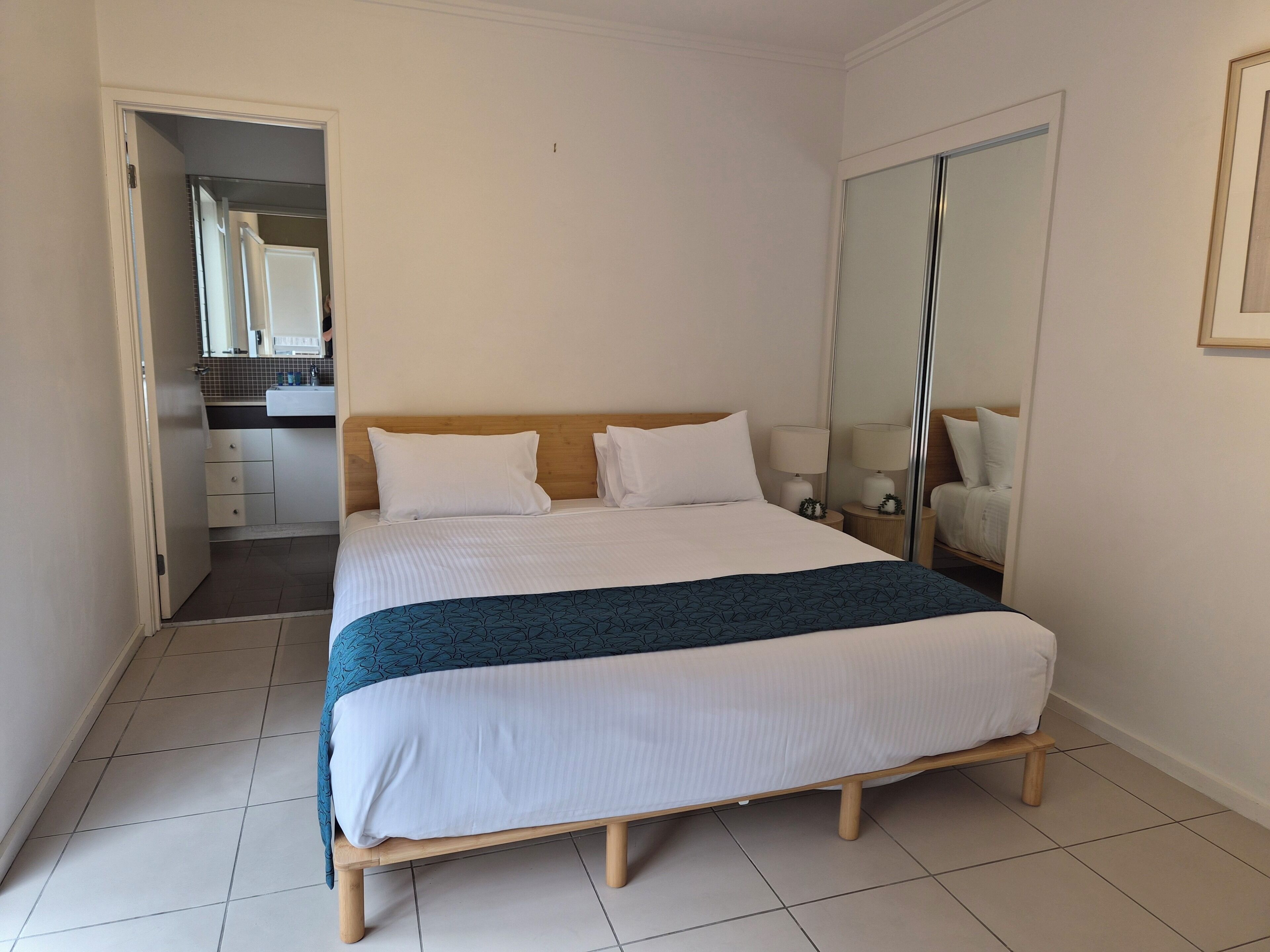A neatly made bed with a wooden frame, a white headboard, and two white pillows. There is a bedside table with a lamp and a small plant. The room has a tiled floor and a large mirror on the wall.