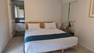 A neatly made bed with a wooden frame, a white headboard, and two white pillows. There is a bedside table with a lamp and a small plant. The room has a tiled floor and a large mirror on the wall.