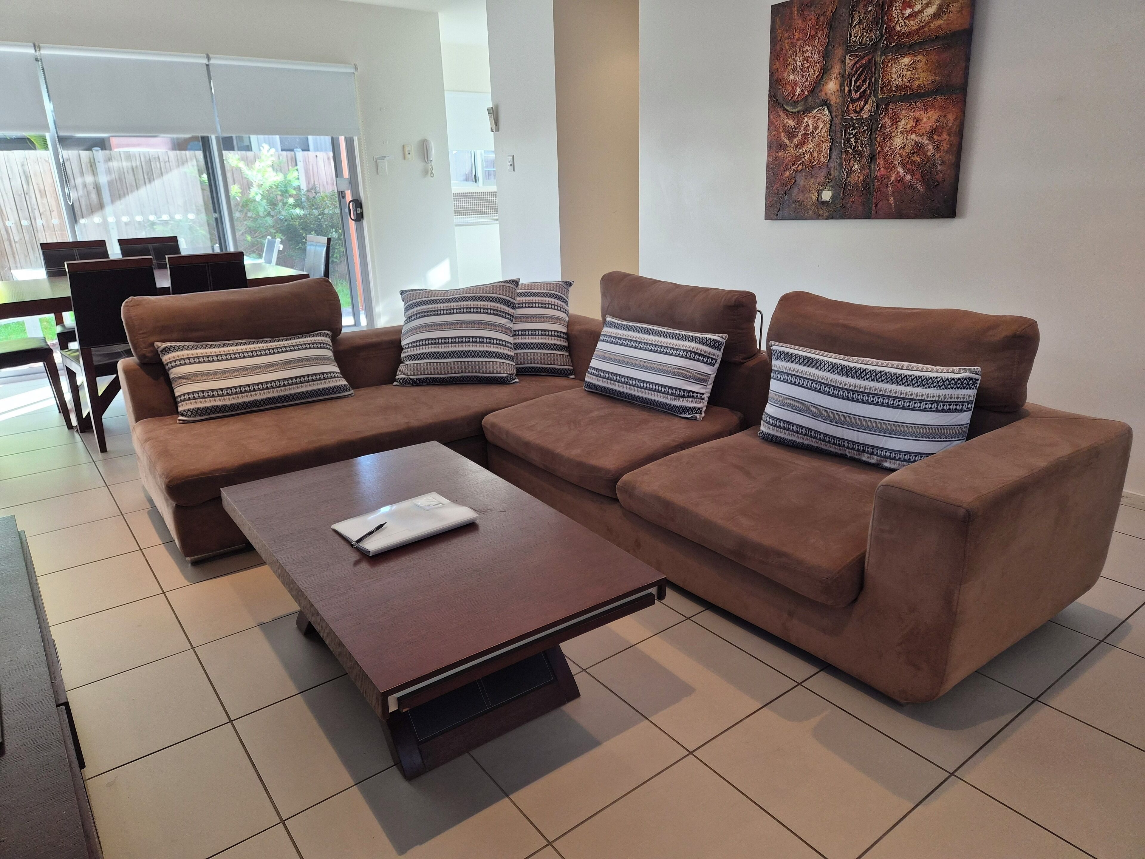 A modern living room with a brown sofa set, patterned cushions, a wooden coffee table, and a large abstract painting on the wall.