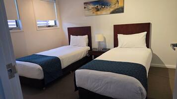 A hotel room with two single beds, a nightstand with a lamp, a painting on the wall, and a window with blinds.
