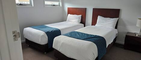 Une chambre d’hôtel avec deux lits simples, une tête de lit en bois, une table de chevet avec une lampe et une fenêtre donnant sur l’extérieur.