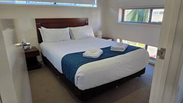 A neatly made bed with white and blue bedding, a bedside table with a lamp, and a window with a view of greenery.