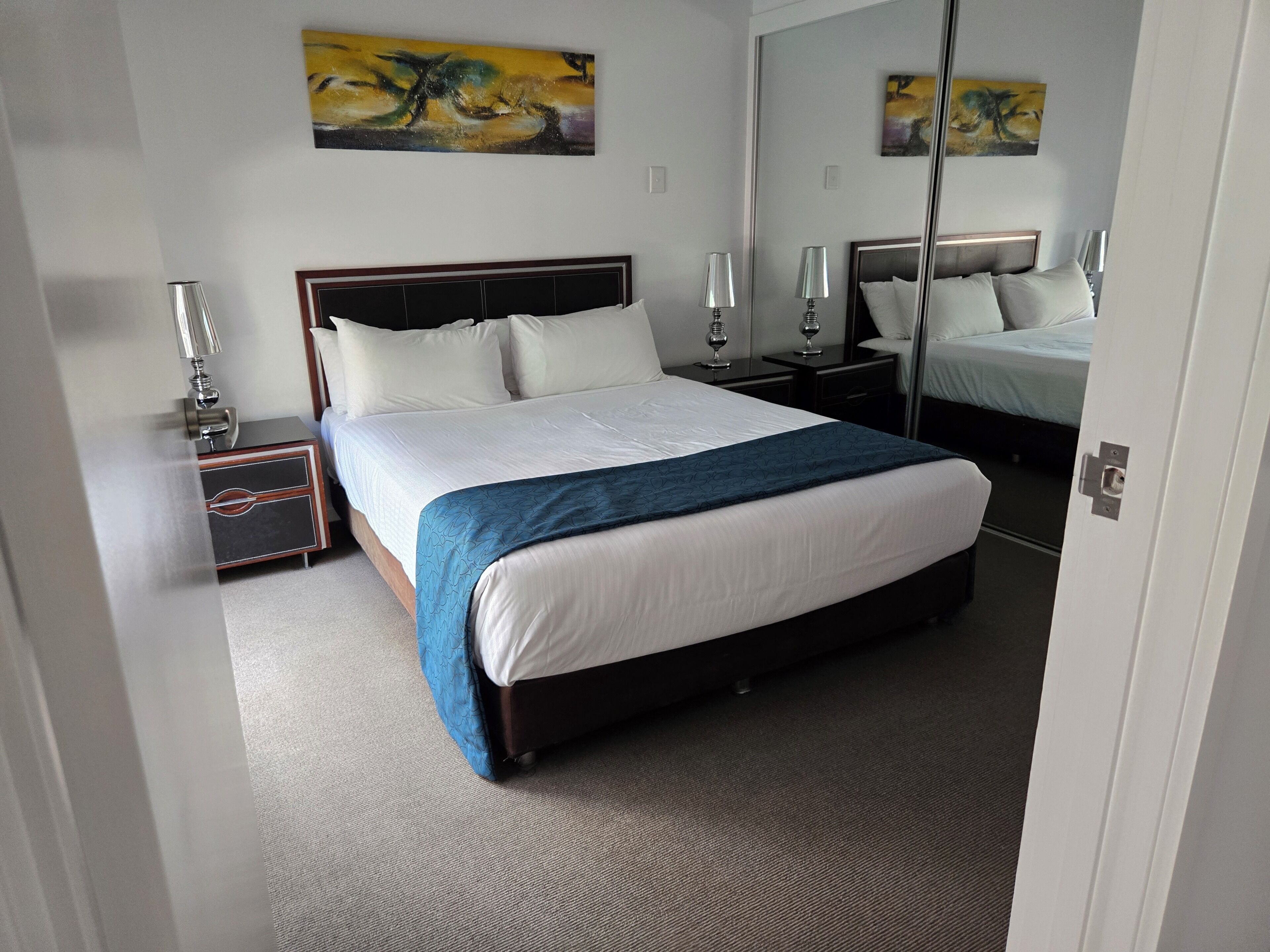 Habitación de hotel con una cama grande, dos mesitas de noche con lámparas, un aparador y dos cuadros abstractos en las paredes.