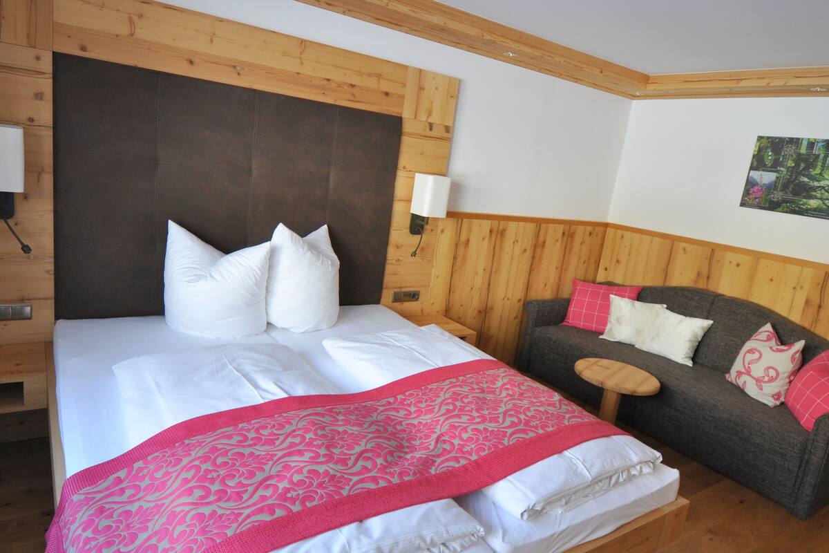 A bedroom with a large bed, a wooden headboard, a grey sofa, and a small wooden table.