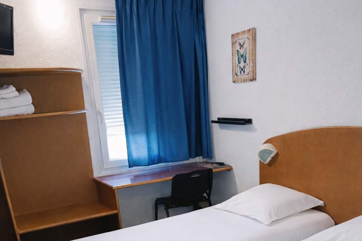 A hotel room with two single beds, a desk with a chair, a TV, and a window with blue curtains.