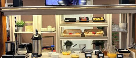 Una barra de desayuno con equipo para preparar café, un televisor mostrando a un grupo de personas y diversos alimentos para el desayuno.