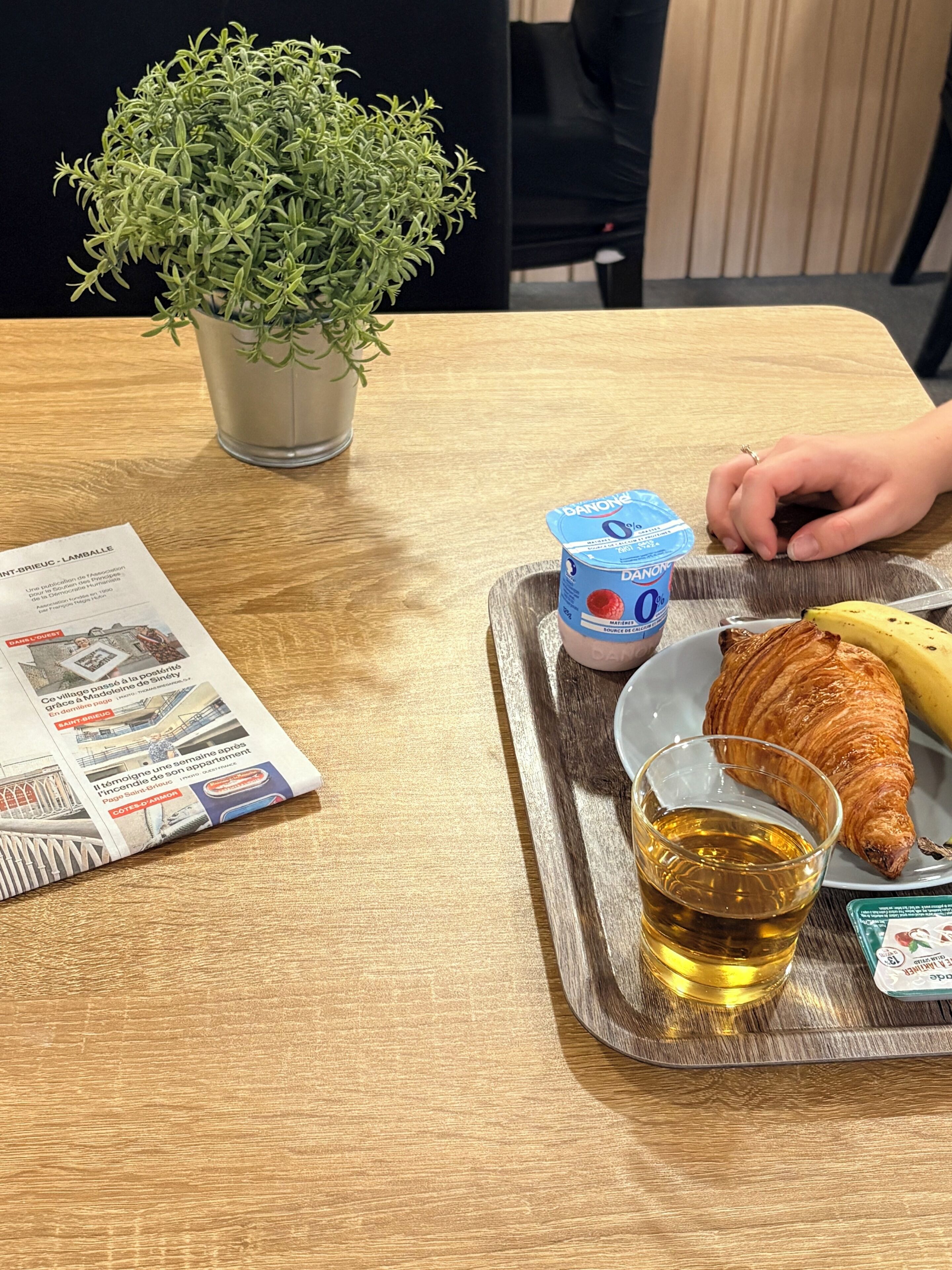 A tray with a croissant, a banana, a glass of amber liquid, and a container of yogurt on a wooden table.
