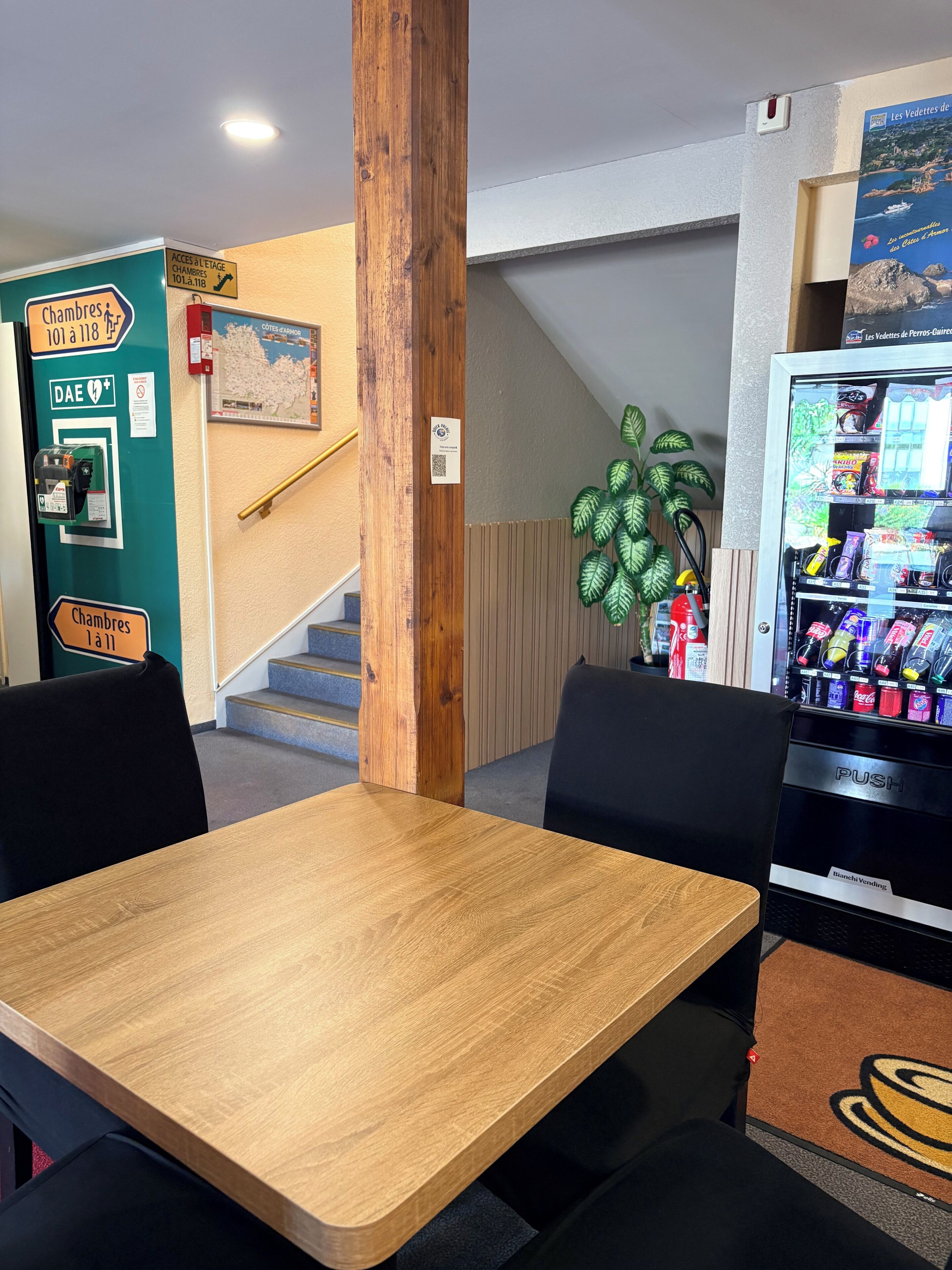 Une table en bois, des chaises noires, un distributeur automatique et un panneau indiquant l’accès à la salle.