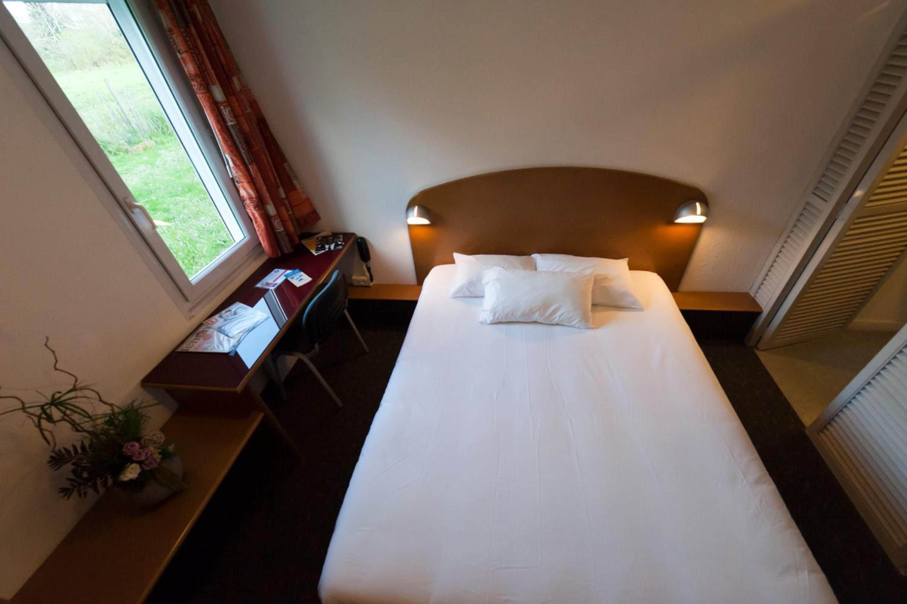 A hotel room with a bed, bedside table, lamp, window with curtains, and a desk with a computer.