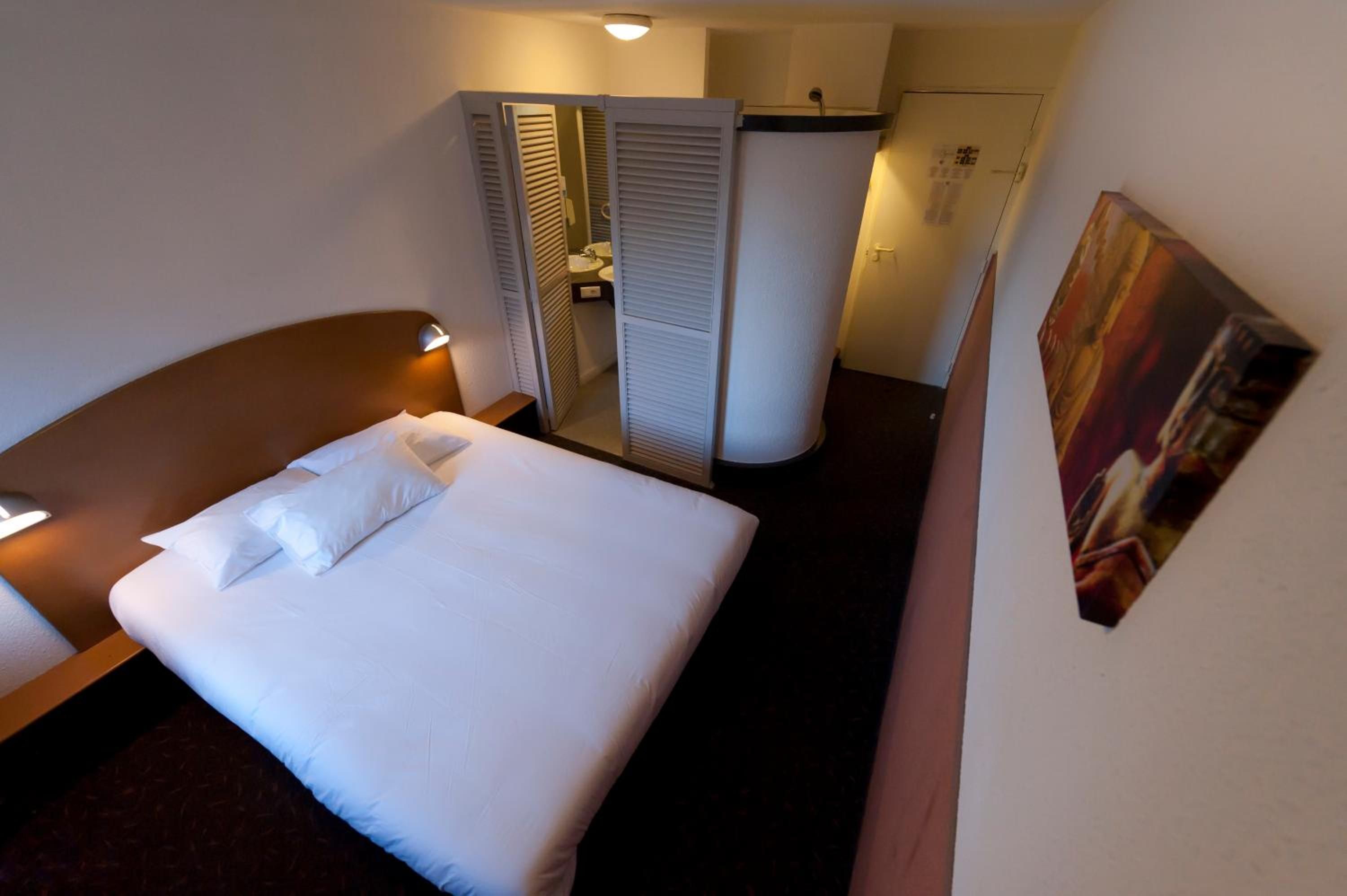 A hotel room with a bed, a bedside table, a wardrobe, and a door leading to a bathroom.