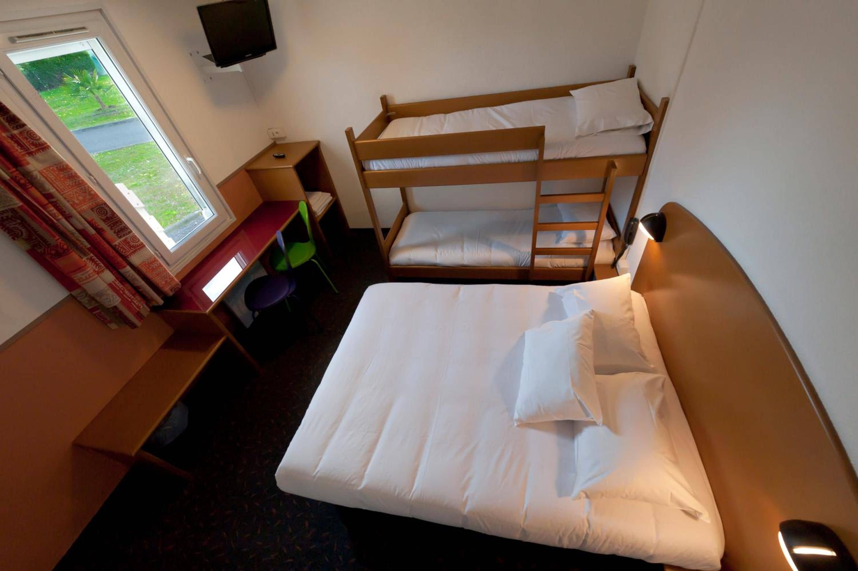Une chambre d’hôtel avec un lit superposé, un bureau avec une chaise, une télévision et une fenêtre avec des rideaux.