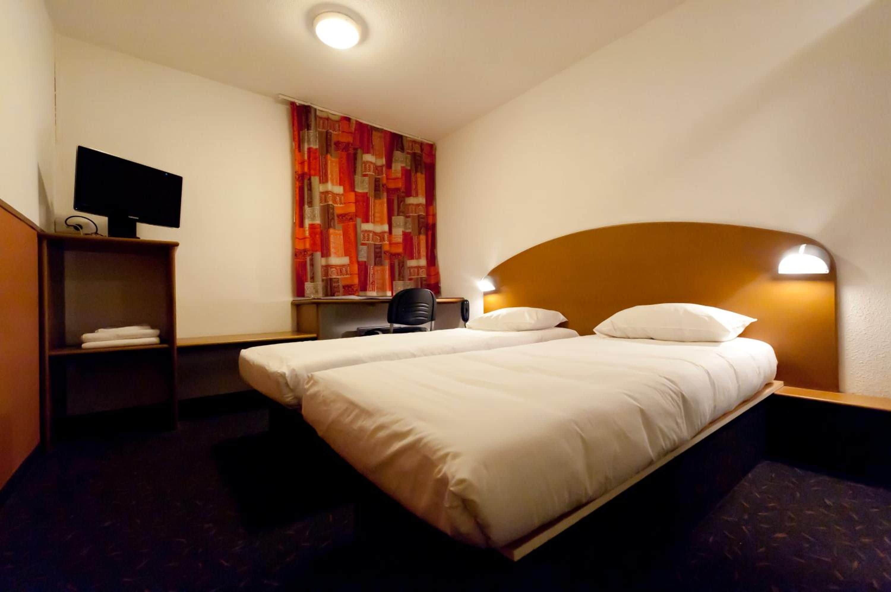 A hotel room with two beds, a desk with a chair, a TV, and a patterned wall.