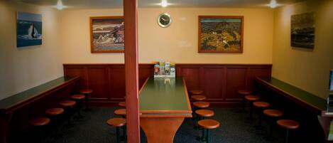 Uma área de bar com uma mesa central e banquetas, cercada por painéis de madeira e obras de arte nas paredes.