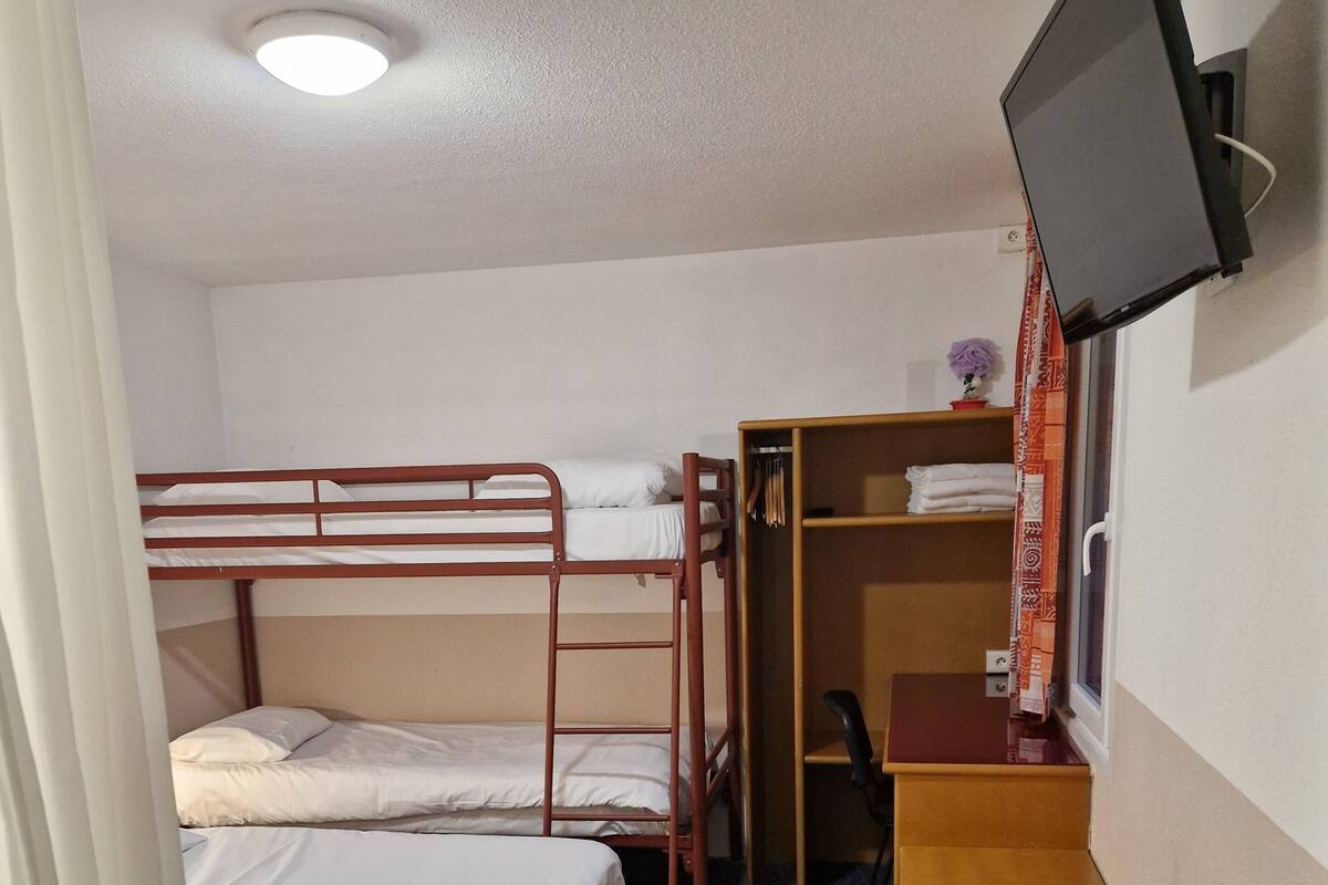 Une chambre d’hôtel compacte équipée de lits superposés, d’un bureau, d’une télévision et d’une petite poubelle.