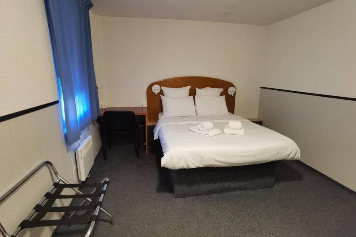 A hotel room with a single bed, a bedside table, a chair, and a window with blue curtains.