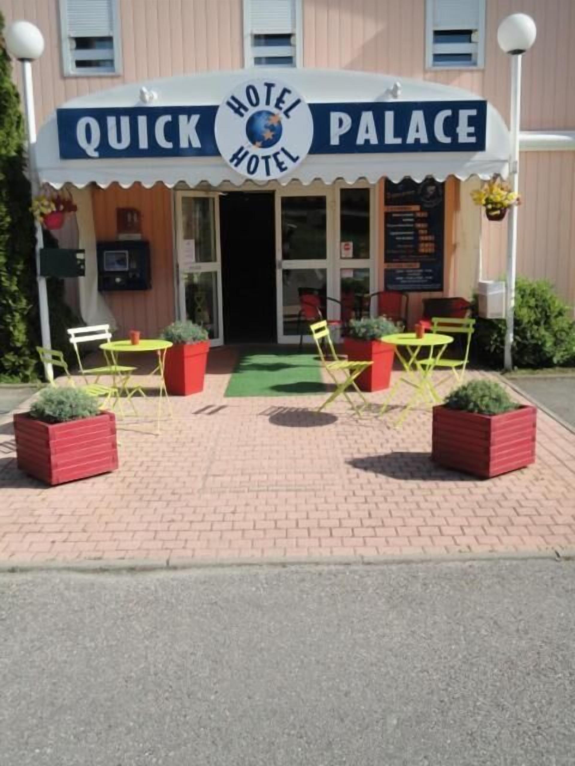 A hotel named "Quick Palace" with a sign above the entrance, outdoor seating with tables and chairs, and potted plants.