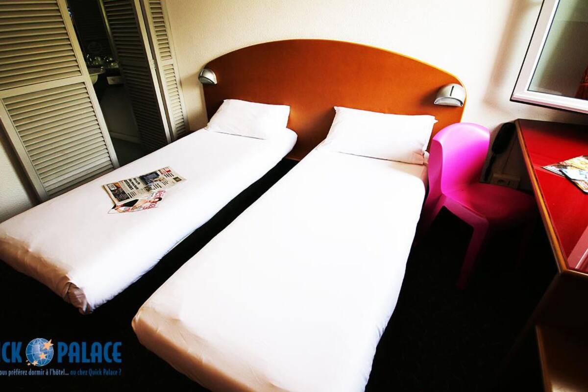A hotel room with two single beds, a wooden headboard, a small desk with a chair, and a television.