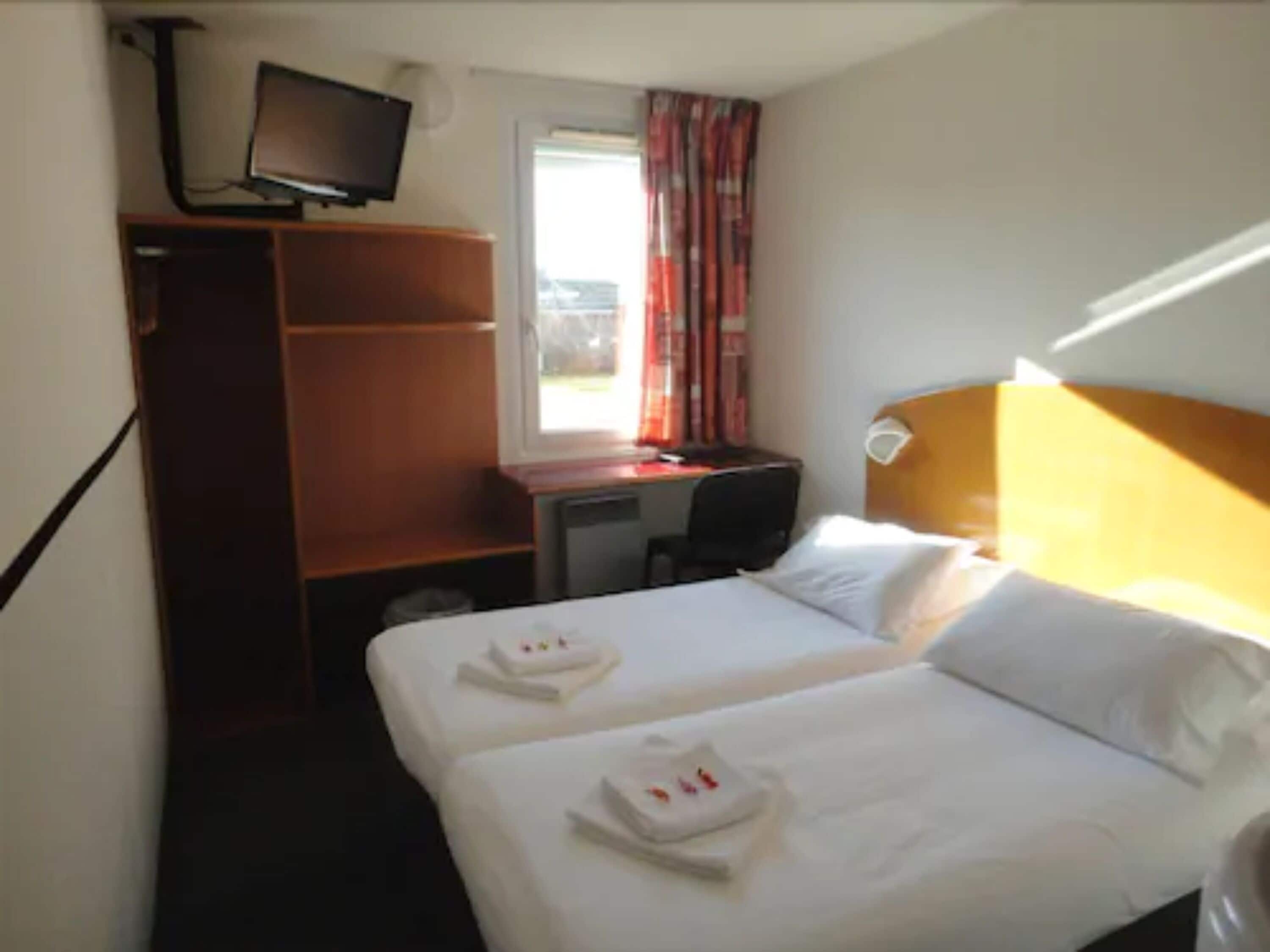 Quarto de hotel com duas camas, uma escrivaninha, uma cadeira, uma televisão e uma janela com cortinas.