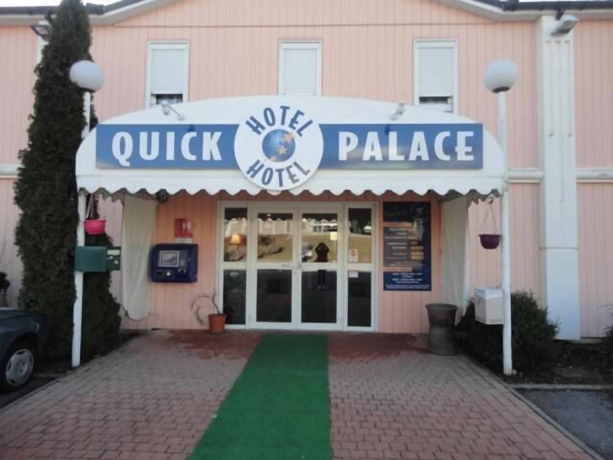 Um hotel chamado "Quick Hotel Palace" com uma placa sobre a entrada, um tapete verde levando à porta e uma caixa de correio à esquerda.