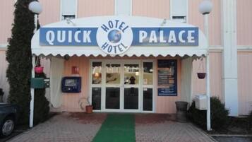 Um hotel chamado "Quick Hotel Palace" com uma placa sobre a entrada, um tapete verde levando à porta e uma caixa de correio à esquerda.