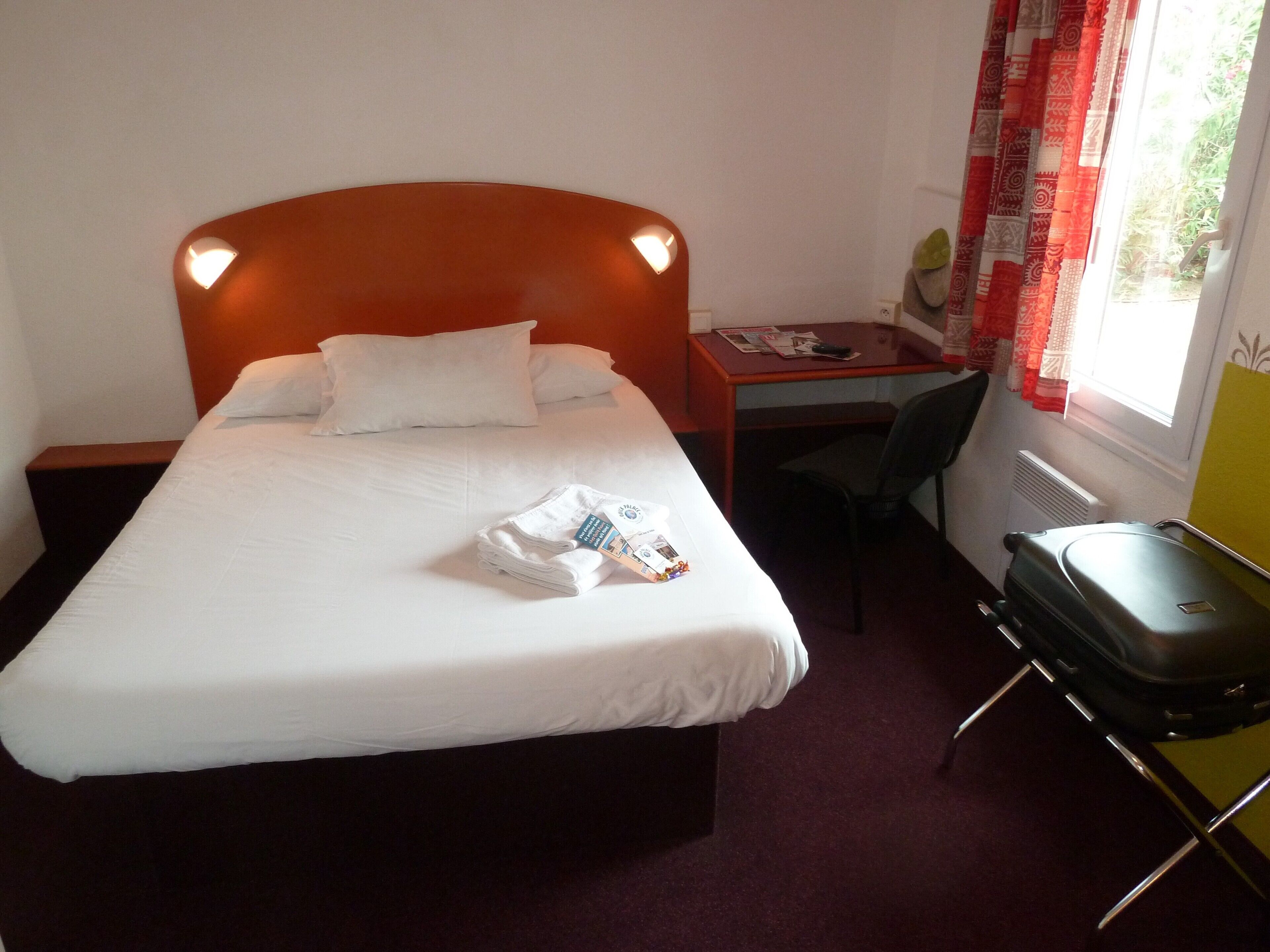 Une chambre d’hôtel avec un lit simple, un bureau, une chaise, une valise et une fenêtre avec des rideaux.