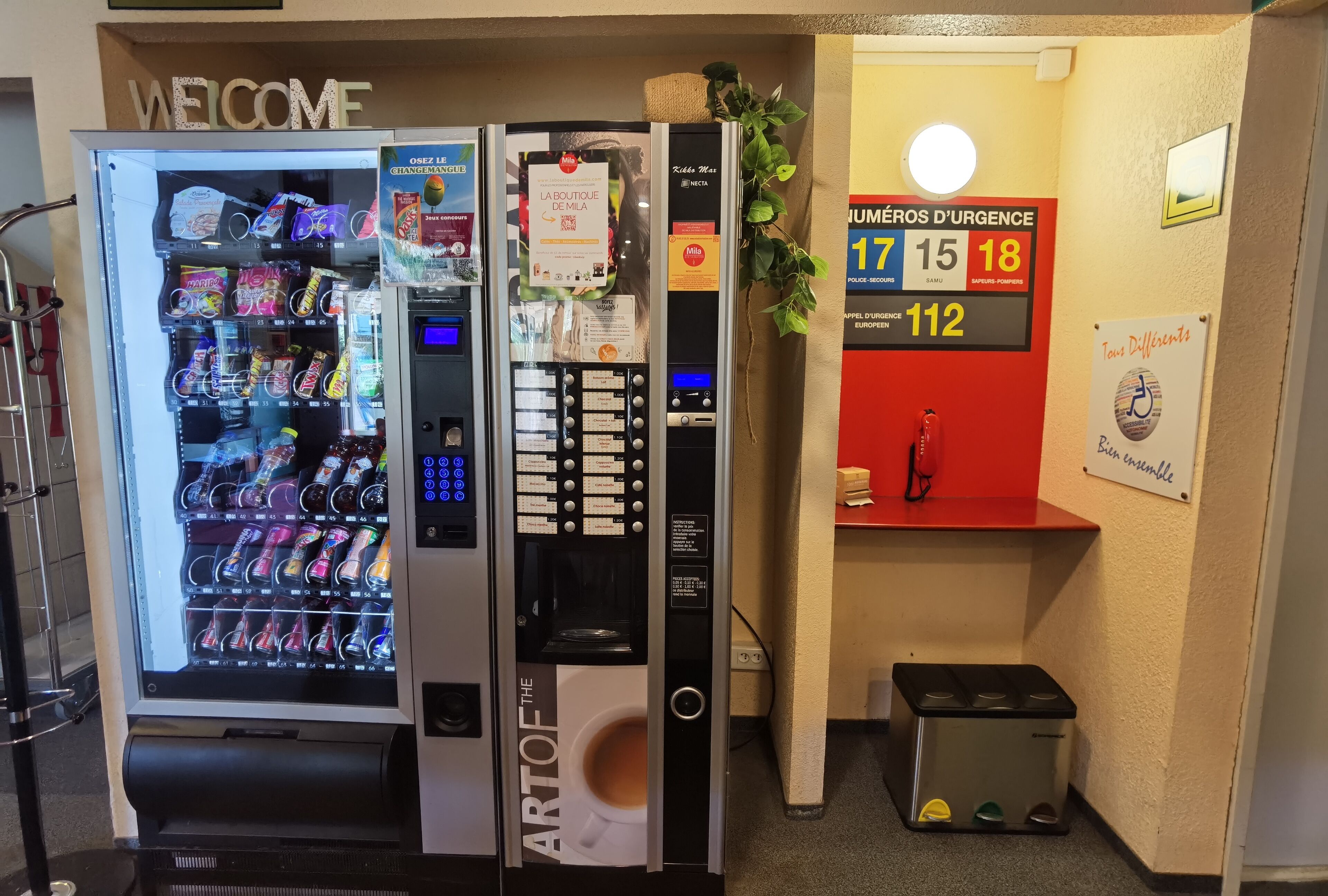 A vending machine with a 'Welcome' sign above it, a small plant, and a fire extinguisher on a shelf.