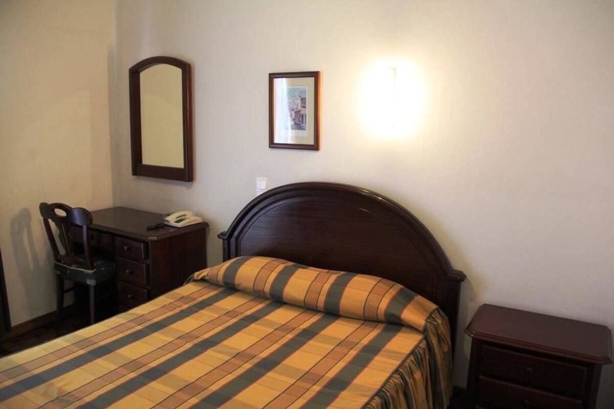 A bedroom with a bed, a desk, a chair, a bedside table, a mirror, and a framed picture on the wall.