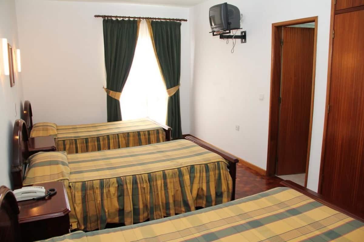 A hotel room with two beds, a television mounted on the wall, a wooden wardrobe, and a window with curtains.